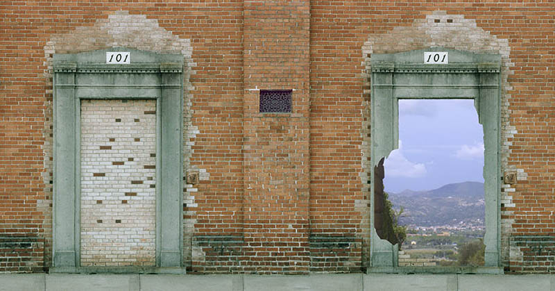 Minibanner: A brick wall with two doors, one blocked by bricks, the other opening onto a blue sky and green hills.